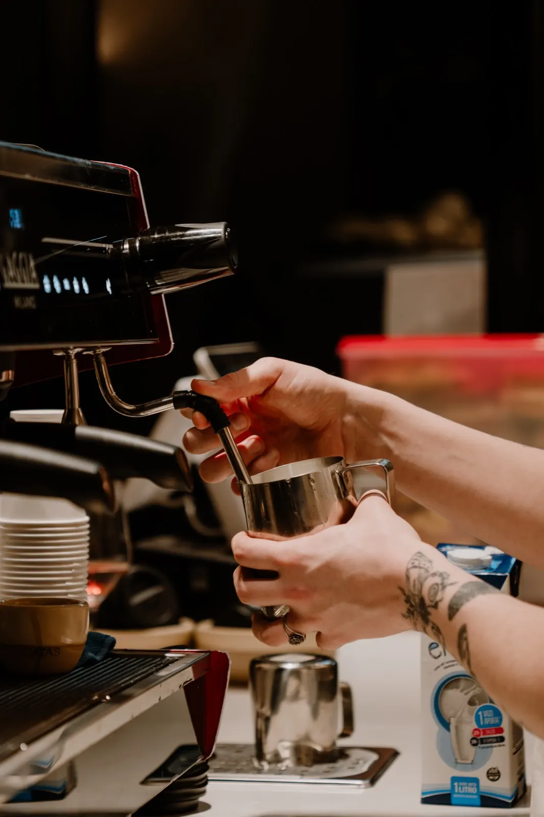Manos de barista que espuman leche en una jarra de acero, junto a la máquina de espresso